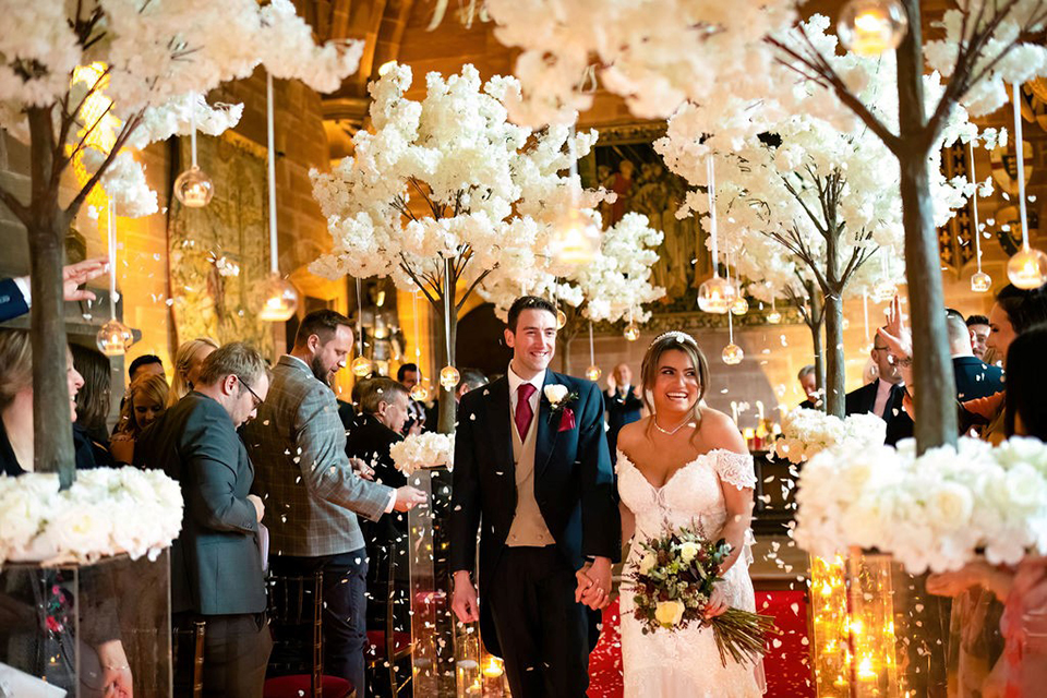 A man wearing a suit and a woman wearing a white wedding dress walking through a brightly lit venue surrounded by white flowers with other people in the background